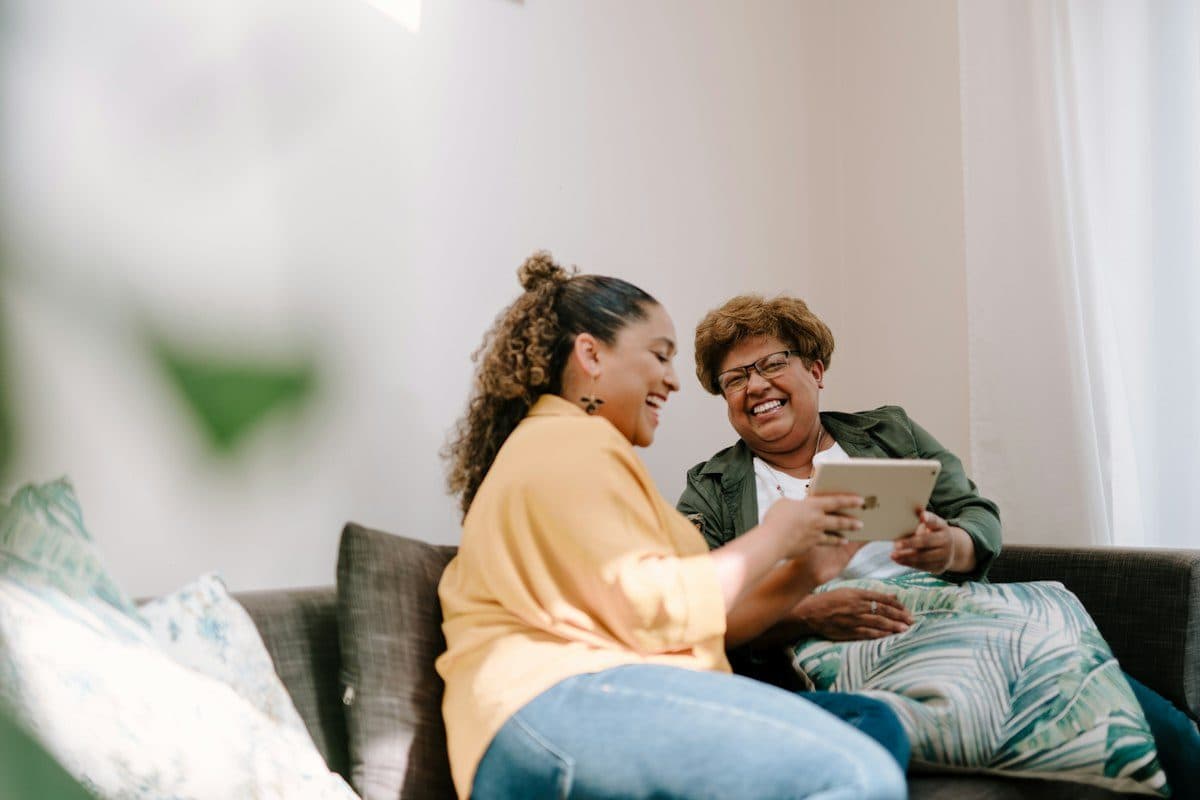 An adult daughter and her father laughing over a tablet on the couch